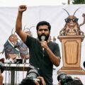 A young South Asian man with a beard speaks passionately into a handheld microphone with his right arm raised in defiance. He stands in front of a large white banner featuring a satirical cartoon mural of cockroaches acting as political figures. To his right, another man stands wearing a black t-shirt and dark sunglasses.