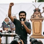 A young South Asian man with a beard speaks passionately into a handheld microphone with his right arm raised in defiance. He stands in front of a large white banner featuring a satirical cartoon mural of cockroaches acting as political figures. To his right, another man stands wearing a black t-shirt and dark sunglasses.