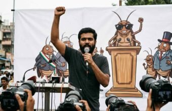 A young South Asian man with a beard speaks passionately into a handheld microphone with his right arm raised in defiance. He stands in front of a large white banner featuring a satirical cartoon mural of cockroaches acting as political figures. To his right, another man stands wearing a black t-shirt and dark sunglasses.