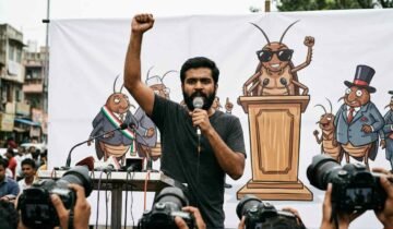 A young South Asian man with a beard speaks passionately into a handheld microphone with his right arm raised in defiance. He stands in front of a large white banner featuring a satirical cartoon mural of cockroaches acting as political figures. To his right, another man stands wearing a black t-shirt and dark sunglasses.