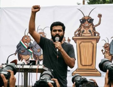 A young South Asian man with a beard speaks passionately into a handheld microphone with his right arm raised in defiance. He stands in front of a large white banner featuring a satirical cartoon mural of cockroaches acting as political figures. To his right, another man stands wearing a black t-shirt and dark sunglasses.