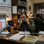 English: A frustrated FIA cybercrime officer in Peshawar sitting at a desk overflowing with unresolved case files and outdated equipment.