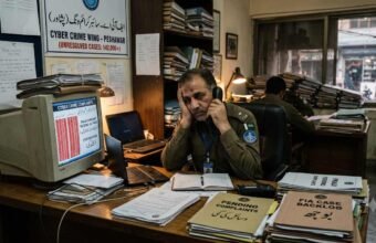 English: A frustrated FIA cybercrime officer in Peshawar sitting at a desk overflowing with unresolved case files and outdated equipment.