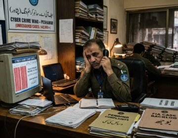 English: A frustrated FIA cybercrime officer in Peshawar sitting at a desk overflowing with unresolved case files and outdated equipment.