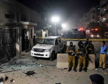 "Crime scene aftermath of the Gulistan-e-Jauhar Rangers attack in Karachi, showing damaged gate, destroyed vehicle, heavy security, and police presence."
