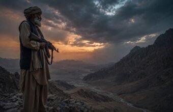 "A gritty, cinematic photojournalism style photograph capturing the tension in the remote tribal districts of Khyber Pakhtunkhwa near the border of Balochistan. A weathered Pashtun elder, a member of a local 'Aman Lashkar' (Peace Committee), is standing vigil on a rocky mountain pass at dusk. He is silhouetted against a dramatic, turbulent twilight sky featuring deep dusty orange and moody blue storm clouds. He is wearing a traditional shalwar kameez and a worn turban, holding an older model Kalashnikov rifle, and looking out over a vast, rugged, desolate mountain valley. The atmosphere is somber, tense, and quiet. The composition emphasizes depth and uncertainty. High contrast, deep shadows, textured film grain, shot on professional editorial camera equipment. Aspect ratio 16:9 (for a website featured image)