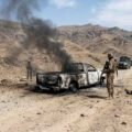 Armed soldiers stand near a heavily burned-out pickup truck with black smoke billowing, on a rugged dirt road in a desert mountain range.