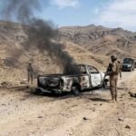Armed soldiers stand near a heavily burned-out pickup truck with black smoke billowing, on a rugged dirt road in a desert mountain range.