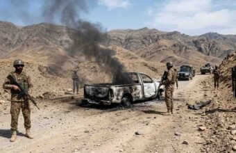 Armed soldiers stand near a heavily burned-out pickup truck with black smoke billowing, on a rugged dirt road in a desert mountain range.