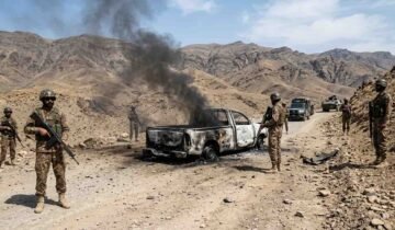Armed soldiers stand near a heavily burned-out pickup truck with black smoke billowing, on a rugged dirt road in a desert mountain range.