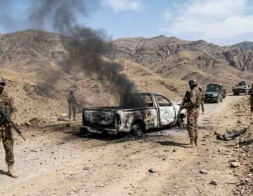 Armed soldiers stand near a heavily burned-out pickup truck with black smoke billowing, on a rugged dirt road in a desert mountain range.