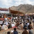 "A wide-angle photograph capturing an 'All Parties Conference' and sit-in held at the foothills of the mountains in the Hanna Urak region of Balochistan. Hundreds of male participants, dressed in traditional Balochi shalwar kameez and turbans, are seated on colorful mats spread on the ground. Behind the stage, a large banner is displayed with the protest's demands written in both Urdu and English: '1. Recovery of 11 Missing Persons', '2. Clearance of Area from Armed Groups', and '3. Talks with Empowered Government Authorities'. A few leaders are standing on the stage, speaking into microphones. Participants are holding placards displaying slogans for the recovery of missing persons. Green and white flags are waving in the air, and an arid mountain range is visible in the background under a cloudy sky."