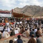 "A wide-angle photograph capturing an 'All Parties Conference' and sit-in held at the foothills of the mountains in the Hanna Urak region of Balochistan. Hundreds of male participants, dressed in traditional Balochi shalwar kameez and turbans, are seated on colorful mats spread on the ground. Behind the stage, a large banner is displayed with the protest's demands written in both Urdu and English: '1. Recovery of 11 Missing Persons', '2. Clearance of Area from Armed Groups', and '3. Talks with Empowered Government Authorities'. A few leaders are standing on the stage, speaking into microphones. Participants are holding placards displaying slogans for the recovery of missing persons. Green and white flags are waving in the air, and an arid mountain range is visible in the background under a cloudy sky."