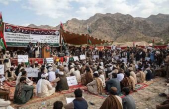 "A wide-angle photograph capturing an 'All Parties Conference' and sit-in held at the foothills of the mountains in the Hanna Urak region of Balochistan. Hundreds of male participants, dressed in traditional Balochi shalwar kameez and turbans, are seated on colorful mats spread on the ground. Behind the stage, a large banner is displayed with the protest's demands written in both Urdu and English: '1. Recovery of 11 Missing Persons', '2. Clearance of Area from Armed Groups', and '3. Talks with Empowered Government Authorities'. A few leaders are standing on the stage, speaking into microphones. Participants are holding placards displaying slogans for the recovery of missing persons. Green and white flags are waving in the air, and an arid mountain range is visible in the background under a cloudy sky."