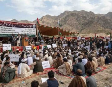 "A wide-angle photograph capturing an 'All Parties Conference' and sit-in held at the foothills of the mountains in the Hanna Urak region of Balochistan. Hundreds of male participants, dressed in traditional Balochi shalwar kameez and turbans, are seated on colorful mats spread on the ground. Behind the stage, a large banner is displayed with the protest's demands written in both Urdu and English: '1. Recovery of 11 Missing Persons', '2. Clearance of Area from Armed Groups', and '3. Talks with Empowered Government Authorities'. A few leaders are standing on the stage, speaking into microphones. Participants are holding placards displaying slogans for the recovery of missing persons. Green and white flags are waving in the air, and an arid mountain range is visible in the background under a cloudy sky."