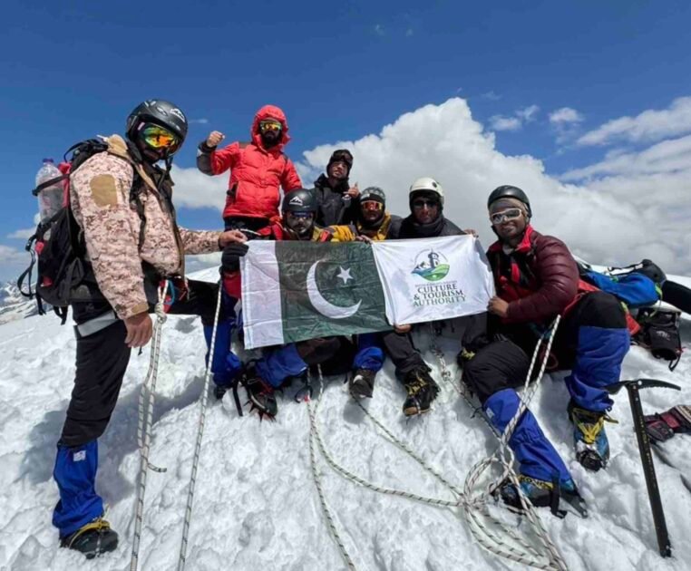 Nine climbers from Khyber Pakhtunkhwa celebrate after summiting the 6,050-meter Thalo Zom Peak, holding the Pakistani flag and the KP Culture & Tourism Authority flag.