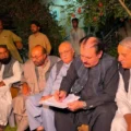 A medium shot of a man in a black waistcoat signing the Ziarat martyrs protest agreement. He is surrounded by other men, including a white-haired elder with glasses, tribal leaders, and government officials from Balochistan. The setting is a garden at night, with a pomegranate tree branch visible.