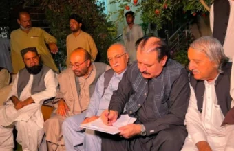 A medium shot of a man in a black waistcoat signing the Ziarat martyrs protest agreement. He is surrounded by other men, including a white-haired elder with glasses, tribal leaders, and government officials from Balochistan. The setting is a garden at night, with a pomegranate tree branch visible.