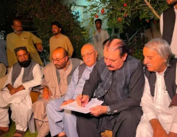 A medium shot of a man in a black waistcoat signing the Ziarat martyrs protest agreement. He is surrounded by other men, including a white-haired elder with glasses, tribal leaders, and government officials from Balochistan. The setting is a garden at night, with a pomegranate tree branch visible.