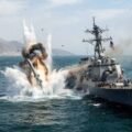 Text: A dramatic explosion erupts in the water directly adjacent to a US Navy Arleigh Burke-class guided-missile destroyer (USS Porter, DDG-78) in the Strait of Hormuz, with rocky coastal mountains visible in the background.