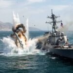 Text: A dramatic explosion erupts in the water directly adjacent to a US Navy Arleigh Burke-class guided-missile destroyer (USS Porter, DDG-78) in the Strait of Hormuz, with rocky coastal mountains visible in the background.
