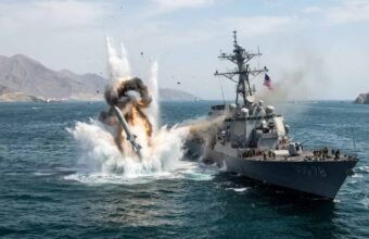 Text: A dramatic explosion erupts in the water directly adjacent to a US Navy Arleigh Burke-class guided-missile destroyer (USS Porter, DDG-78) in the Strait of Hormuz, with rocky coastal mountains visible in the background.
