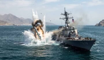 Text: A dramatic explosion erupts in the water directly adjacent to a US Navy Arleigh Burke-class guided-missile destroyer (USS Porter, DDG-78) in the Strait of Hormuz, with rocky coastal mountains visible in the background.