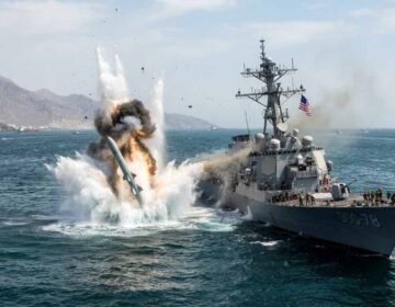 Text: A dramatic explosion erupts in the water directly adjacent to a US Navy Arleigh Burke-class guided-missile destroyer (USS Porter, DDG-78) in the Strait of Hormuz, with rocky coastal mountains visible in the background.