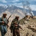 A photojournalist's photograph shows four armed Afghan men in traditional clothing and tactical gear walking along a rocky mountain path. They carry rifles and an RPG. In the background are snow-capped mountains, a village, and smoke rising from a small, damaged fort flying a white Taliban flag. Watermark "THE KHYBER TIMES".