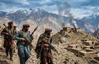 A photojournalist's photograph shows four armed Afghan men in traditional clothing and tactical gear walking along a rocky mountain path. They carry rifles and an RPG. In the background are snow-capped mountains, a village, and smoke rising from a small, damaged fort flying a white Taliban flag. Watermark "THE KHYBER TIMES".