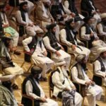 Vertical photograph showing two bearded Afghan men in traditional white clothing, turbans, and black vests sitting in chairs at a formal gathering. The man on the left has a brown patterned turban and holds a pen to his chin thoughtfully. The man on the right has a prominent white beard, a white turban, holds a tablet, and has a checkered shawl over his shoulder. Other similarly dressed men are seated in the background. White text in Urdu script is at the bottom right.
