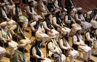 Vertical photograph showing two bearded Afghan men in traditional white clothing, turbans, and black vests sitting in chairs at a formal gathering. The man on the left has a brown patterned turban and holds a pen to his chin thoughtfully. The man on the right has a prominent white beard, a white turban, holds a tablet, and has a checkered shawl over his shoulder. Other similarly dressed men are seated in the background. White text in Urdu script is at the bottom right.