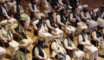 Vertical photograph showing two bearded Afghan men in traditional white clothing, turbans, and black vests sitting in chairs at a formal gathering. The man on the left has a brown patterned turban and holds a pen to his chin thoughtfully. The man on the right has a prominent white beard, a white turban, holds a tablet, and has a checkered shawl over his shoulder. Other similarly dressed men are seated in the background. White text in Urdu script is at the bottom right.