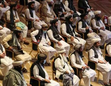 Vertical photograph showing two bearded Afghan men in traditional white clothing, turbans, and black vests sitting in chairs at a formal gathering. The man on the left has a brown patterned turban and holds a pen to his chin thoughtfully. The man on the right has a prominent white beard, a white turban, holds a tablet, and has a checkered shawl over his shoulder. Other similarly dressed men are seated in the background. White text in Urdu script is at the bottom right.