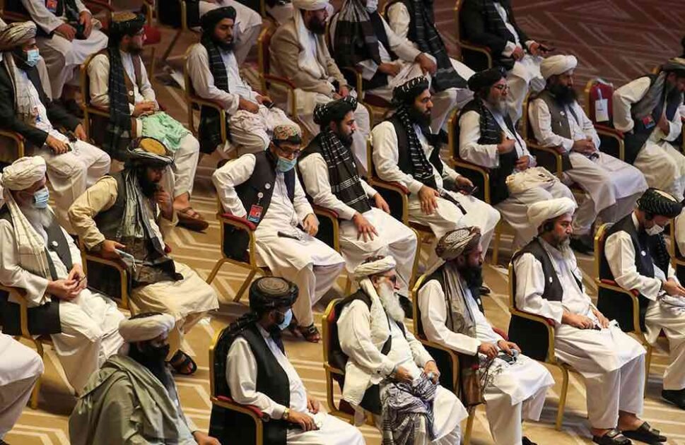 Vertical photograph showing two bearded Afghan men in traditional white clothing, turbans, and black vests sitting in chairs at a formal gathering. The man on the left has a brown patterned turban and holds a pen to his chin thoughtfully. The man on the right has a prominent white beard, a white turban, holds a tablet, and has a checkered shawl over his shoulder. Other similarly dressed men are seated in the background. White text in Urdu script is at the bottom right.