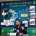 A man with a beard, wearing a dark blazer over a black shirt, is at a press desk with multiple microphones, gesturing with his hand. Behind him is a large screen with a detailed infographic. The main title on the screen is in Urdu and English: "بھارتی ڈس انفارمیشن نیٹ ورک بے نقاب: فنڈنگ کا پردہ چاک" (Indian Disinformation Network Exposed: Funding Revealed) and "پاکستانی حساس اداروں کا کامیاب آپریشن" (Successful Operation by Pakistani Sensitive Agencies). The infographic visualizes a flow of Indian Rupee coins and notes from a map of India on the left ('Indian Source') towards a grid of interconnected social media icons (Facebook, X, YouTube, TikTok, Telegram) on the right, labeled 'Disinformation Network.' Arrows indicate the flow of funding and misinformation. The screen also includes three panels with examples of social media posts labeled 'Fake News' related to the AJK Joint Action Committee, with images of conflict. Text explicitly labels these as 'Fake' and 'جعلی'.