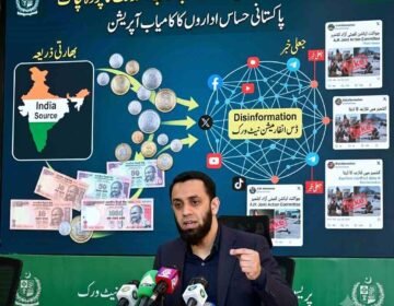A man with a beard, wearing a dark blazer over a black shirt, is at a press desk with multiple microphones, gesturing with his hand. Behind him is a large screen with a detailed infographic. The main title on the screen is in Urdu and English: "بھارتی ڈس انفارمیشن نیٹ ورک بے نقاب: فنڈنگ کا پردہ چاک" (Indian Disinformation Network Exposed: Funding Revealed) and "پاکستانی حساس اداروں کا کامیاب آپریشن" (Successful Operation by Pakistani Sensitive Agencies). The infographic visualizes a flow of Indian Rupee coins and notes from a map of India on the left ('Indian Source') towards a grid of interconnected social media icons (Facebook, X, YouTube, TikTok, Telegram) on the right, labeled 'Disinformation Network.' Arrows indicate the flow of funding and misinformation. The screen also includes three panels with examples of social media posts labeled 'Fake News' related to the AJK Joint Action Committee, with images of conflict. Text explicitly labels these as 'Fake' and 'جعلی'.