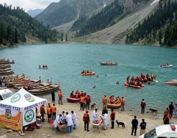 Rescue 1122 emergency response operation at an alpine lake in Swat Valley, showing an overturned tourist boat, rescue inflatable rafts, ambulances, and a medical tent on the shore.