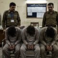 An image of three handcuffed men sitting with their heads bowed in a police station investigation room, flanked by two uniformed police officers.
