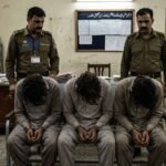 An image of three handcuffed men sitting with their heads bowed in a police station investigation room, flanked by two uniformed police officers.