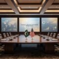 "A luxurious, empty conference room set up for the Pakistan-US Strategic Trade Dialogue in Washington D.C., featuring a wooden table with the flags of Pakistan and the United States, and large digital screens displaying global economic data."