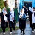 Four young Afghan schoolgirls in black uniforms and white headscarves walking together on a street in Kabul.