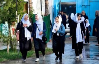 Four young Afghan schoolgirls in black uniforms and white headscarves walking together on a street in Kabul.