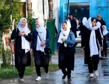 Four young Afghan schoolgirls in black uniforms and white headscarves walking together on a street in Kabul.