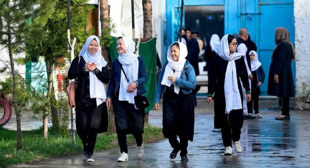 Four young Afghan schoolgirls in black uniforms and white headscarves walking together on a street in Kabul.
