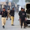 Pakistani security personnel from KP Police and Army standing on a dusty urban street in Peshawar, Khyber Pakhtunkhwa, next to a police vehicle with license plate CH 2645. Buildings with Urdu signs are visible.