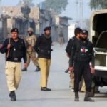 Pakistani security personnel from KP Police and Army standing on a dusty urban street in Peshawar, Khyber Pakhtunkhwa, next to a police vehicle with license plate CH 2645. Buildings with Urdu signs are visible.
