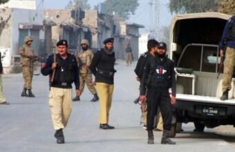 Pakistani security personnel from KP Police and Army standing on a dusty urban street in Peshawar, Khyber Pakhtunkhwa, next to a police vehicle with license plate CH 2645. Buildings with Urdu signs are visible.