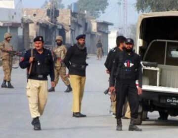 Pakistani security personnel from KP Police and Army standing on a dusty urban street in Peshawar, Khyber Pakhtunkhwa, next to a police vehicle with license plate CH 2645. Buildings with Urdu signs are visible.