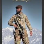 NRF field commander Haseeb Panjshiri standing on a mountain peak in military camouflage gear and a pakol hat holding a rifle against snow-capped Afghan mountains.